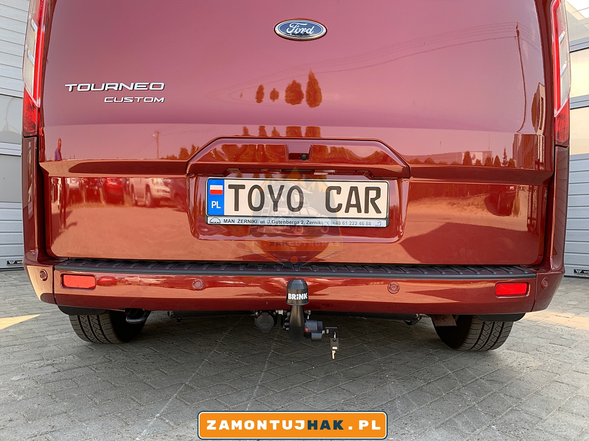 Zdjęcie przedstawia Forda Tourneo Custom z zamontowanym hakiem holowniczym wypinanym pionowo brink, zdjęcie zostało zrobione od tyłu ze zbliżeniem na kulę haka holowniczego, na tle serwisu zamontujhak.pl